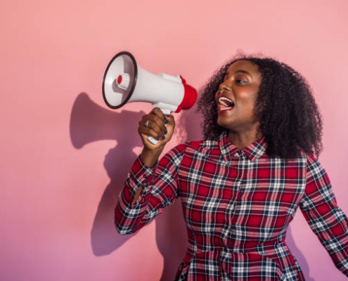 Young black woman standing in front of pink background turns head to side yelling into a megaphone.