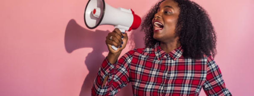 Young black woman standing in front of pink background turns head to side yelling into a megaphone.
