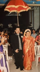 Bridegroom in a black suit and the bride in a red saree with jasmines and firecracker flowers on her hair standing under a red velvet umbrella in a wedding hall. Guests on either side of the couple are clapping their hands. A live band is seen in the background.