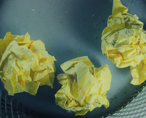 Three crumpled up yellow pieces of paper with some visible but illegible writing, in a waste basket.