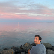 Jonathan Focht sitting on a rock next to the water with a beautiful sky