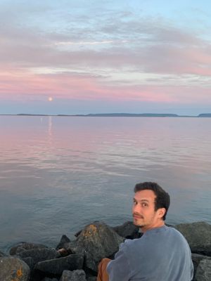 Jonathan Focht sitting on a rock next to the water with a beautiful sky