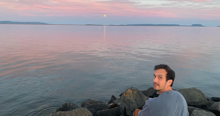 Jonathan Focht sitting on a rock next to the water with a beautiful sky